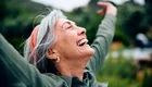 Eine ältere Frau mit grauem Haar und einem orangenen Haarband lächelt glücklich, während sie ihre Arme weit ausbreitet. Ihr Gesicht strahlt Freude und Zufriedenheit aus. Der Hintergrund zeigt eine grüne, natürliche Umgebung.