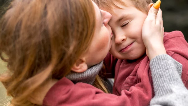 Eine Mutter umarmt ihren kleinen Sohn liebevoll und küsst ihn auf die Wange. Beide wirken glücklich und zufrieden. Die Szene vermittelt eine warme, liebevolle Beziehung zwischen Eltern und Kind in einer natürlichen Umgebung.