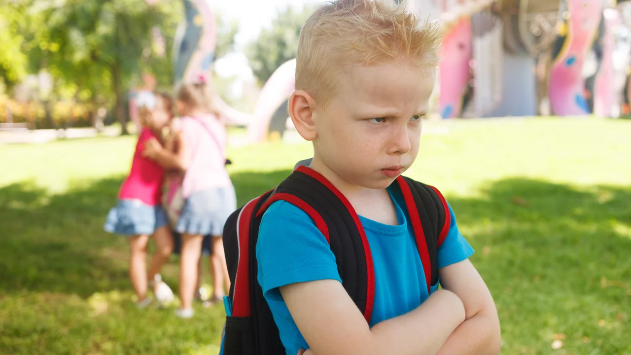Ein unglücklicher Junge mit blondem, kurz geschnittenem Haar und einem Rucksack steht im Vordergrund. Er hat die Arme vor der Brust verschränkt und schaut verärgert. Im Hintergrund sind drei andere Kinder, die gemeinsam spielen und lachen.