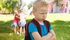 Ein unglücklicher Junge mit blondem, kurz geschnittenem Haar und einem Rucksack steht im Vordergrund. Er hat die Arme vor der Brust verschränkt und schaut verärgert. Im Hintergrund sind drei andere Kinder, die gemeinsam spielen und lachen.