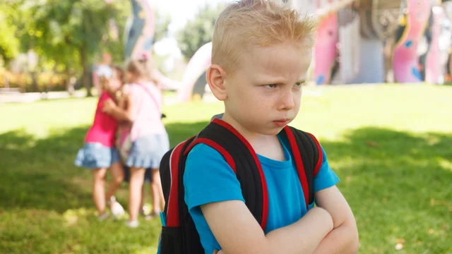 Ein unglücklicher Junge mit blondem, kurz geschnittenem Haar und einem Rucksack steht im Vordergrund. Er hat die Arme vor der Brust verschränkt und schaut verärgert. Im Hintergrund sind drei andere Kinder, die gemeinsam spielen und lachen.