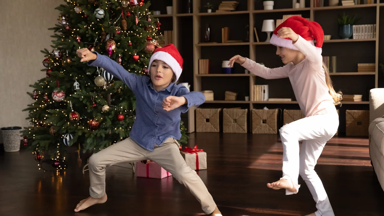 Zwei Kinder tanzen fröhlich vor einem geschmückten Weihnachtsbaum. Sie tragen rote Weihnachtsmützen und lächeln, während sie sich in fröhlichen Posen bewegen. Geschenke liegen unter dem Baum, was die festliche Stimmung unterstreicht.