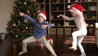 Zwei Kinder tanzen fröhlich vor einem geschmückten Weihnachtsbaum. Sie tragen rote Weihnachtsmützen und lächeln, während sie sich in fröhlichen Posen bewegen. Geschenke liegen unter dem Baum, was die festliche Stimmung unterstreicht.