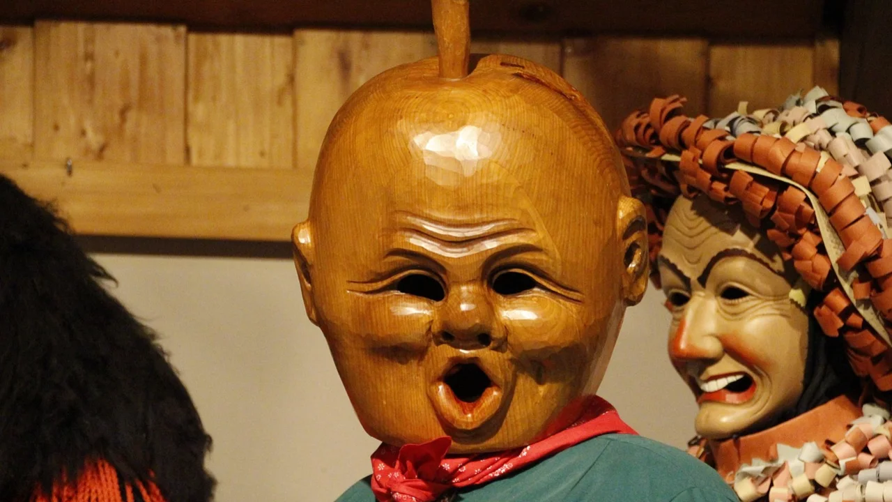 Das Bild zeigt zwei traditionelle Masken aus Holz. Eine Maske hat ein hölzernes, rundes Gesicht mit einem offenen Mund und einer auffälligen Frisur. Die andere Maske lächelt, hat eine komplexe Gestaltung und ist mit einer dekorativen Kopfbedeckung versehen. Beides deutet auf kulturelle Traditionen hin.