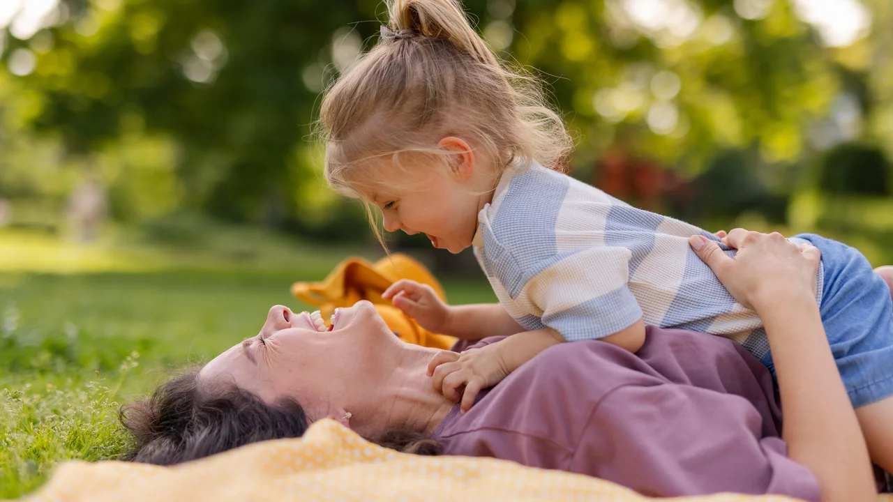 Eine lachende Mutter liegt im Gras und wird von ihrer fröhlichen Tochter, die auf ihr sitzt, umarmt. Beide strahlen Freude und Nähe aus. Im Hintergrund sind verschwommen grüne Bäume zu sehen, was eine entspannte, freundliche Atmosphäre schafft.