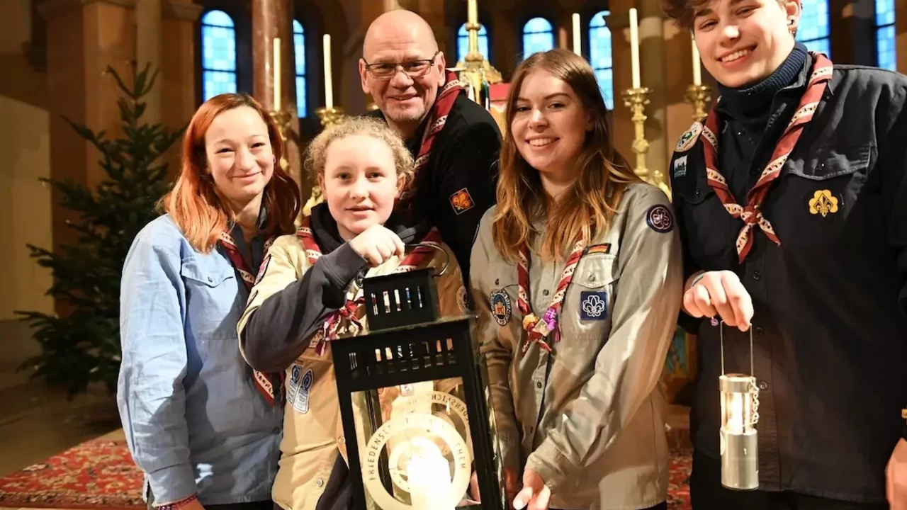 Fünf Personen stehen in einem Innenraum mit einem Weihnachtsbaum im Hintergrund. Sie halten Laternen und tragen Pfadfinderkleidung. Die Gruppe lächelt und posiert zusammen, was auf eine festliche Zusammenkunft hinweist.