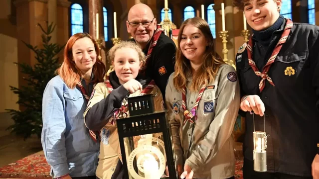 Fünf Personen stehen in einem Innenraum mit einem Weihnachtsbaum im Hintergrund. Sie halten Laternen und tragen Pfadfinderkleidung. Die Gruppe lächelt und posiert zusammen, was auf eine festliche Zusammenkunft hinweist.