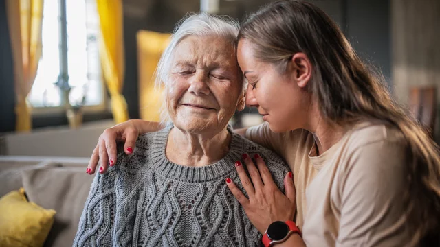 Eine jüngere Frau mit langen braunen Haaren sitzt zusammen mit einer älteren grauhaarigen Frau auf einem Sofa und hat den linken Arm um ihre Schulter und den Kopf an die Stirn der alten Dame gelegt. Die rechte Hand liegt vor der Schulter der alten Dame. Beide haben die Augen geschlossen. Das Bild zeigt eine innige Verbindung zwischen den Beiden.