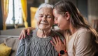 Eine jüngere Frau mit langen braunen Haaren sitzt zusammen mit einer älteren grauhaarigen Frau auf einem Sofa und hat den linken Arm um ihre Schulter und den Kopf an die Stirn der alten Dame gelegt. Die rechte Hand liegt vor der Schulter der alten Dame. Beide haben die Augen geschlossen. Das Bild zeigt eine innige Verbindung zwischen den Beiden.