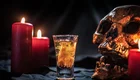 Ein Glas mit einem gelben Getränk steht vor einem Schädel, umgeben von roten Kerzen. Die Kerzen scheinen im Dunkeln zu leuchten und erzeugen eine mystische Atmosphäre.
