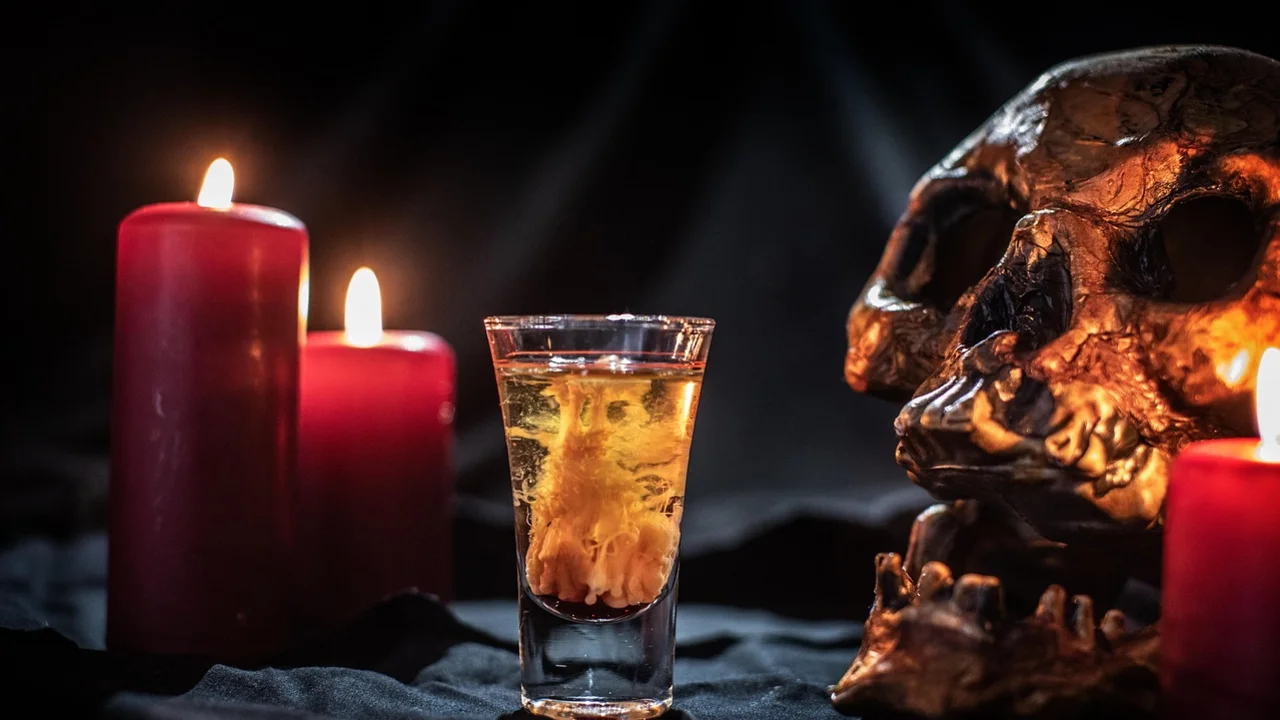 Ein Glas mit einem gelben Getränk steht vor einem Schädel, umgeben von roten Kerzen. Die Kerzen scheinen im Dunkeln zu leuchten und erzeugen eine mystische Atmosphäre.