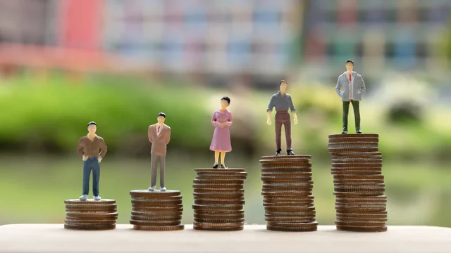 Auf dem Bild stehen fünf kleine Figuren auf verschiedenen Münzhaufen, die unterschiedlich hoch sind. Sie repräsentieren eine Vielfalt von Personen und zeigen eine Aufwärtsentwicklung oder den Vergleich von Werten. Der Hintergrund ist unscharf und zeigt eine grüne Umgebung.