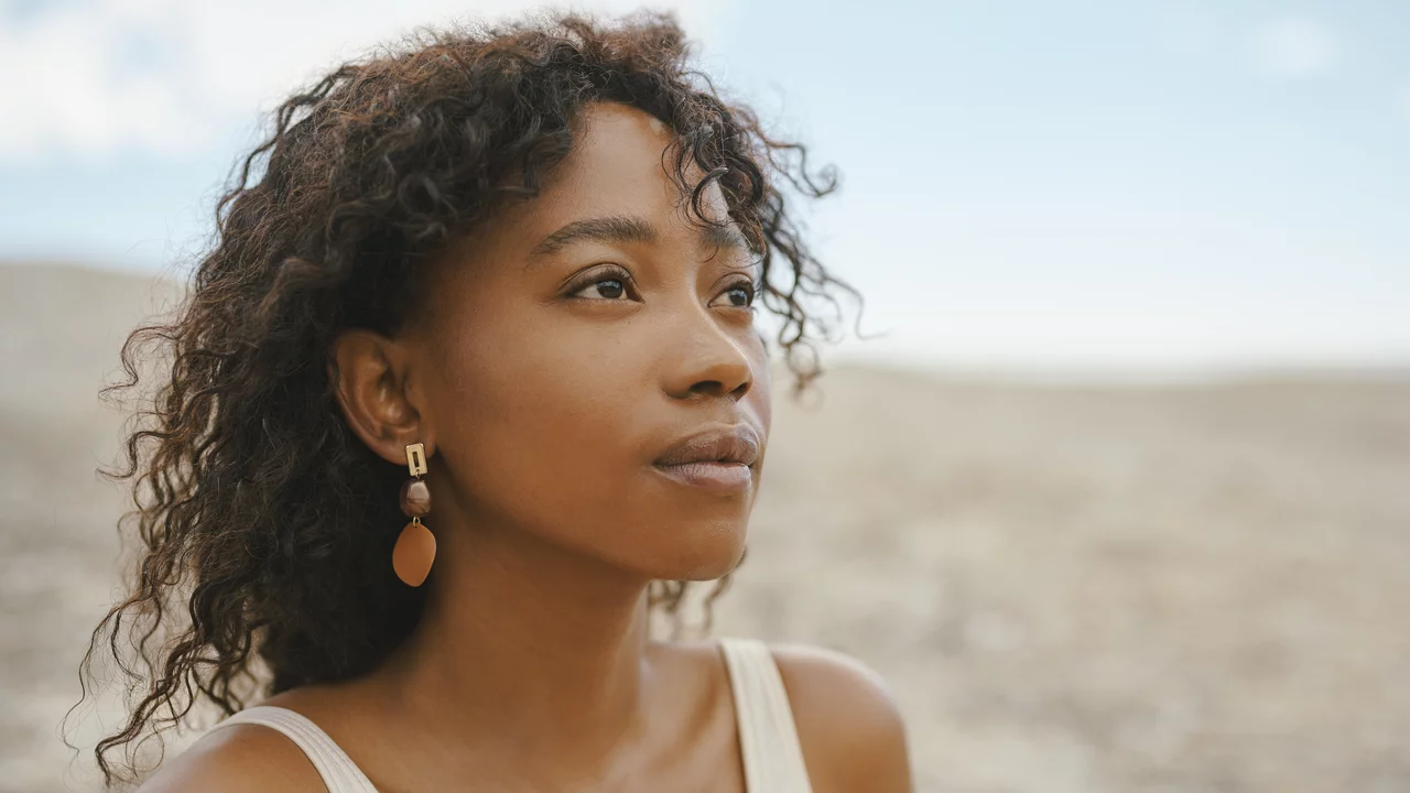 Das Bild zeigt eine Frau mit lockigem Haar, die nachdenklich in die Ferne blickt. Sie trägtOhrringe und ein ärmelloses Oberteil. Der Hintergrund ist unscharf dargestellt, was den Fokus auf ihr Gesicht und ihren Ausdruck legt.