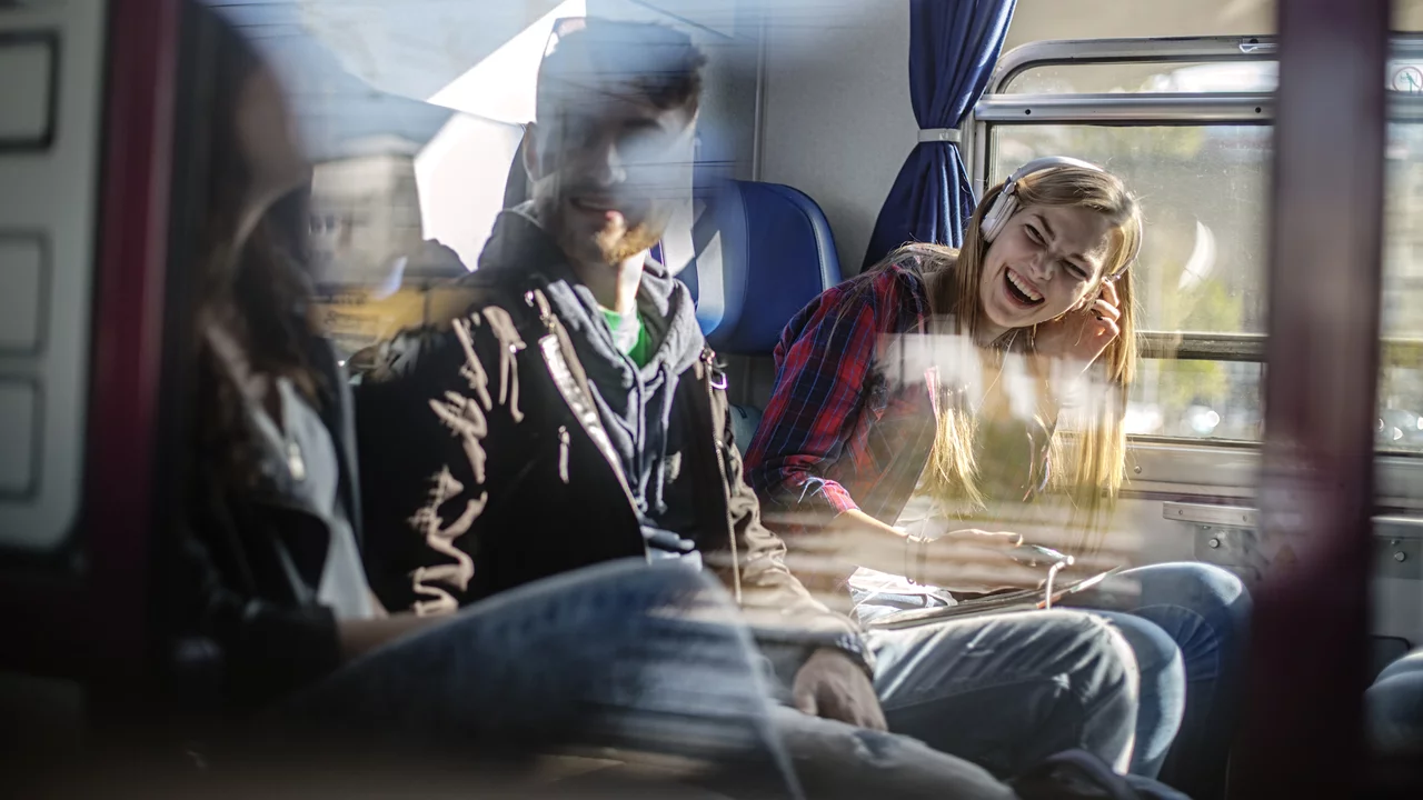 Eine Gruppe von drei jungen Menschen sitzt in einem Zug und lacht miteinander. Der Raum ist hell erleuchtet, wobei einige Reflexionen das Bild leicht verschwommen erscheinen lassen. Die Stimmung wirkt freundlich und entspannt.