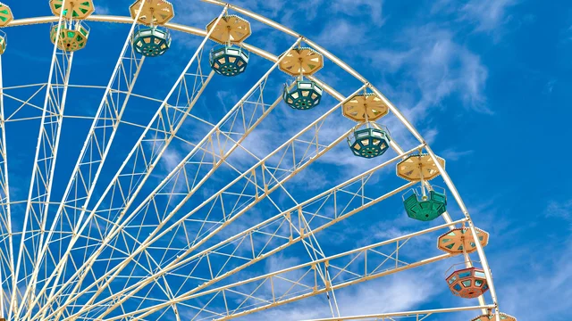 Ein großes Riesenrad mit bunten Gondeln vor einem klaren blauen Himmel. Die Struktur des Riesenrads ist gut sichtbar, wobei die Speichen und das Netz aus Metallrahmen hervortreten.