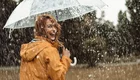 Eine lächelnde Frau mit roten Haaren trägt einen gelben Regenmantel und hält einen durchsichtigen Regenschirm. Sie steht im Regen, während Wasservorhänge um sie herum fallen, und wirkt fröhlich in der herbstlichen, nassen Umgebung.