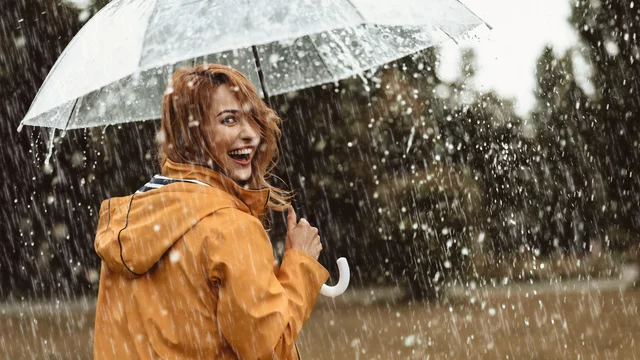 Eine lächelnde Frau mit roten Haaren trägt einen gelben Regenmantel und hält einen durchsichtigen Regenschirm. Sie steht im Regen, während Wasservorhänge um sie herum fallen, und wirkt fröhlich in der herbstlichen, nassen Umgebung.