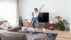 Eine Person mit Kopfhörern tanzt fröhlich und saugt den Boden in einem modernen Wohnzimmer. Im Hintergrund steht ein fernseher und verschiedene Pflanzen sind im Raum verteilt. Die Atmosphäre ist lebhaft und freundlich.
