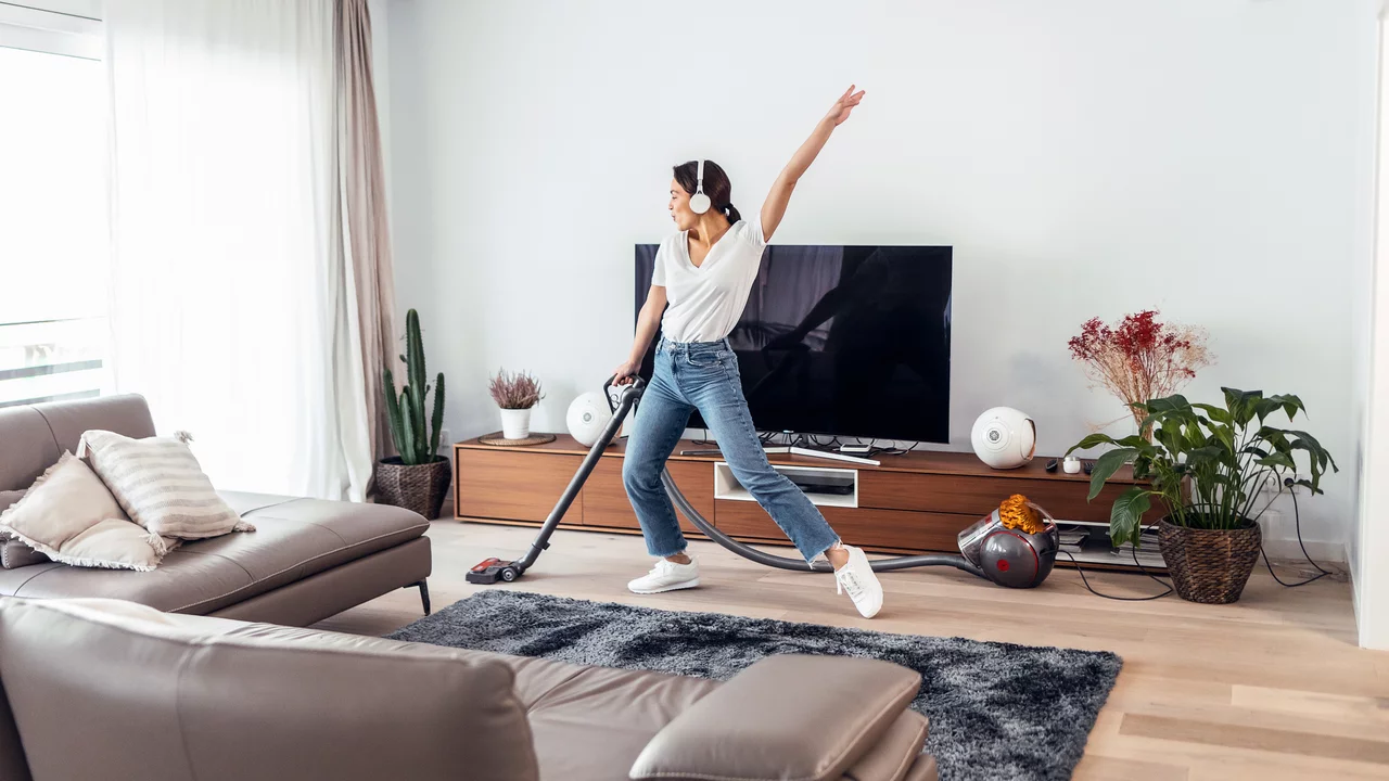 Eine Person mit Kopfhörern tanzt fröhlich und saugt den Boden in einem modernen Wohnzimmer. Im Hintergrund steht ein fernseher und verschiedene Pflanzen sind im Raum verteilt. Die Atmosphäre ist lebhaft und freundlich.