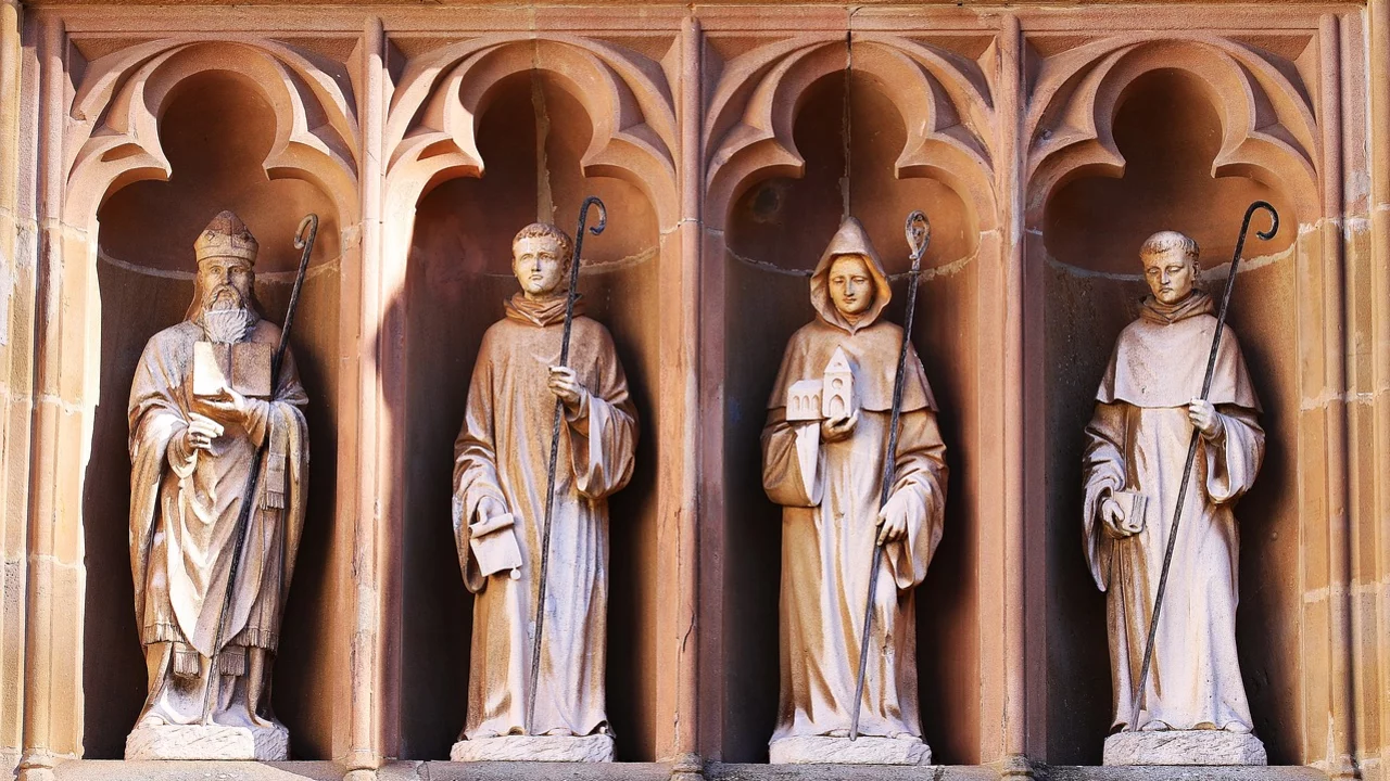 Vier skulptierte Heilige stehen in einem Nischenportal. Jeder trägt charakteristische Gewänder und hält verschiedene Symbole, wie einen Hirtenstab. Die Steinskulpturen sind detailliert gearbeitet und reflektieren religiöse Themen, umgeben von einem architektonischen Rahmen.