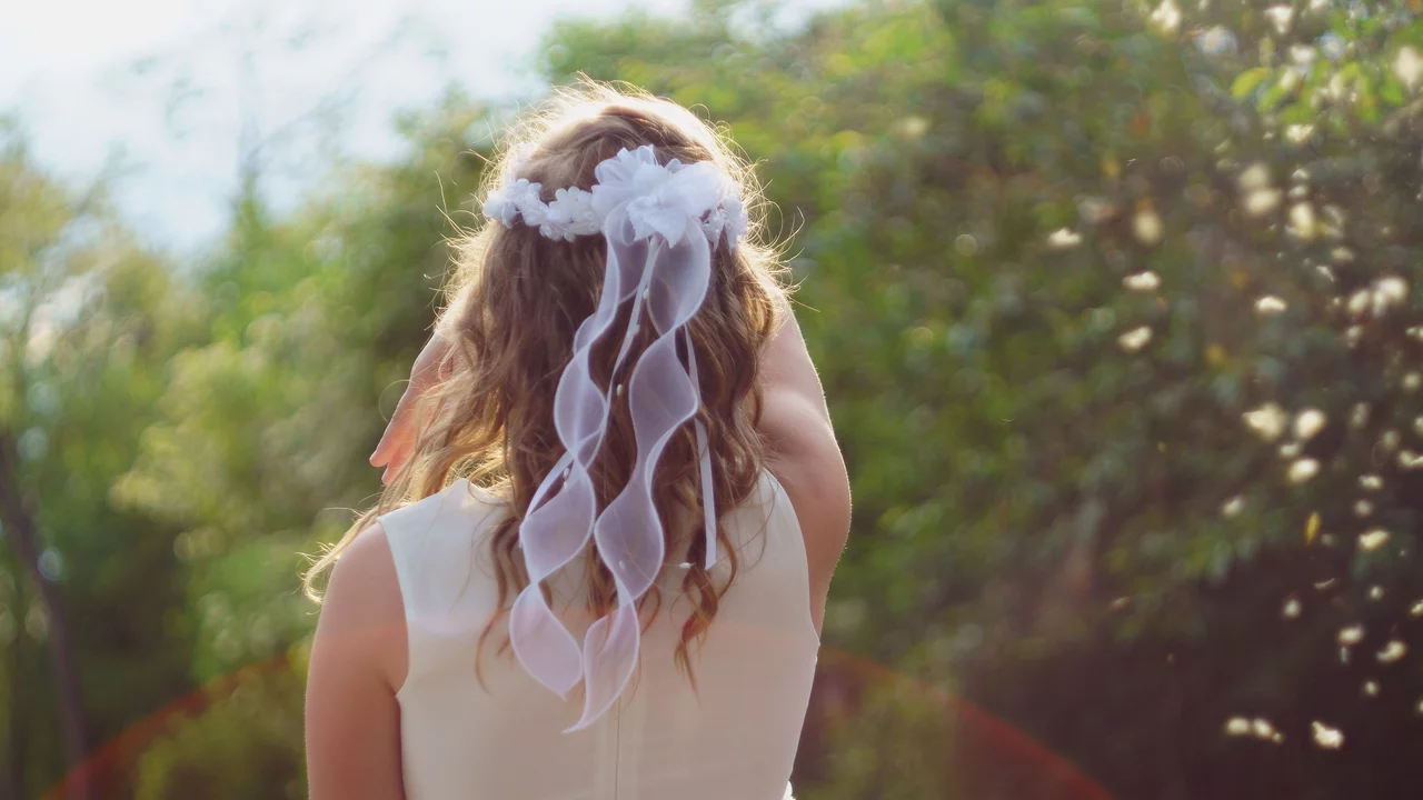 Eine Person mit lockigem, braunem Haar trägt ein weißes Kleid und einen Haarschmuck aus Blumen und Bändern. Sie ist von hinten zu sehen und schaut auf eine grüne, lichtdurchflutete Landschaft.