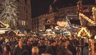 Ein geschäftiger Weihnachtsmarkt bei Nacht, umgeben von festlich beleuchteten Ständen und einem großen Weihnachtsbaum. Viele Menschen schlendern durch die Menge, während die Atmosphäre mit Lichtern und festlicher Stimmung gefüllt ist. Weihnachtsdekorationen sind sichtbar und schaffen eine einladende Stimmung.