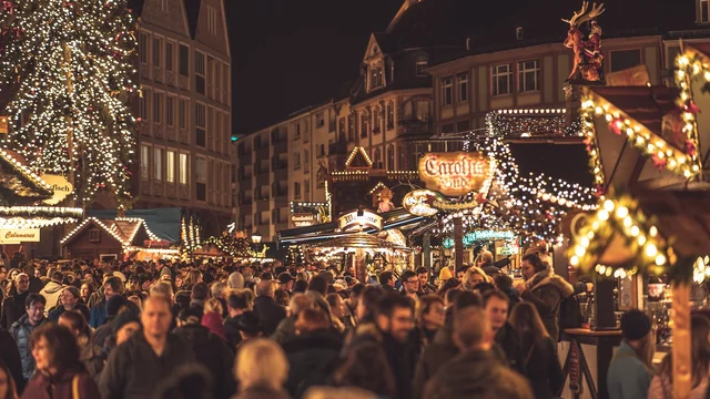 Ein geschäftiger Weihnachtsmarkt bei Nacht, umgeben von festlich beleuchteten Ständen und einem großen Weihnachtsbaum. Viele Menschen schlendern durch die Menge, während die Atmosphäre mit Lichtern und festlicher Stimmung gefüllt ist. Weihnachtsdekorationen sind sichtbar und schaffen eine einladende Stimmung.