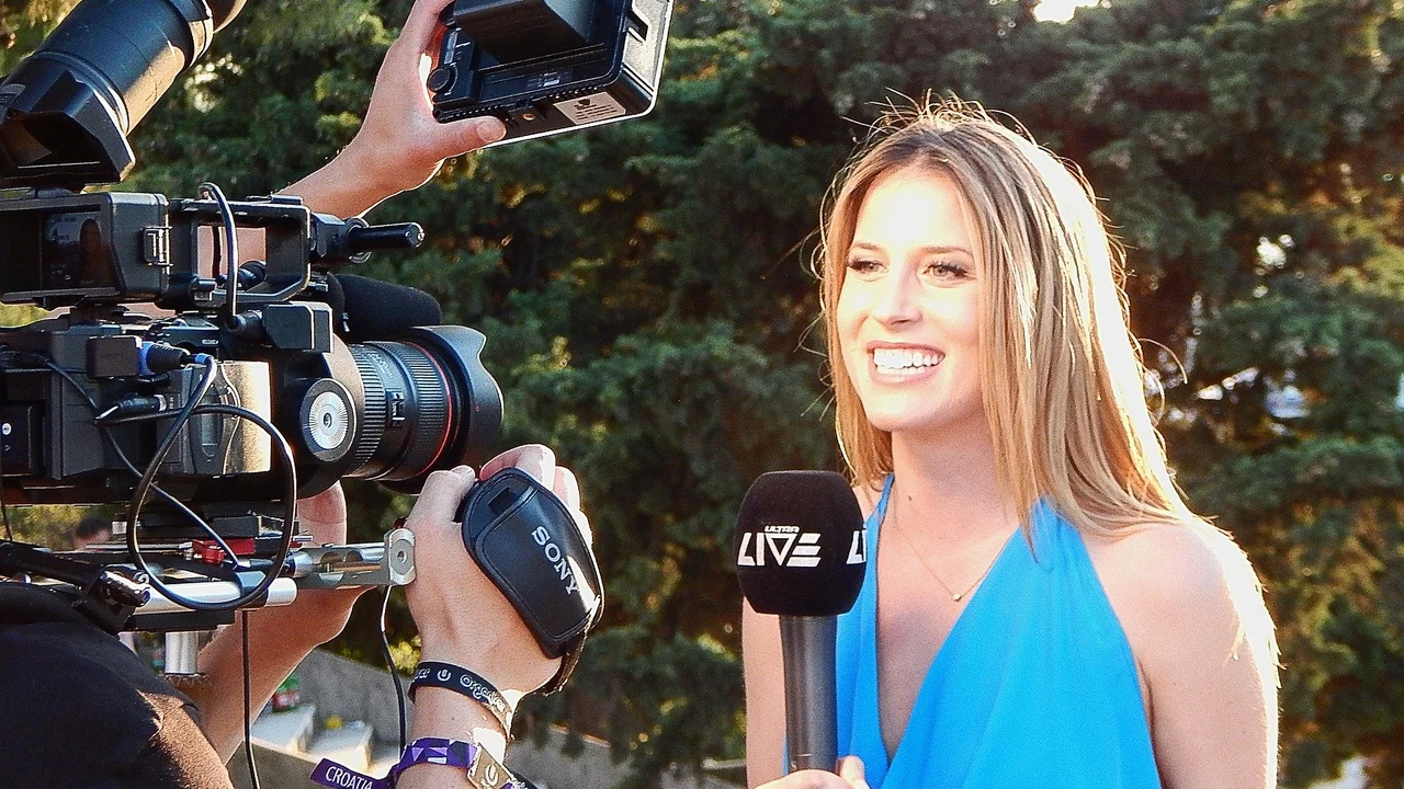 Eine lächelnde Frau in einem blauen Kleid steht vor einer Kamera und spricht in ein Mikrofon. Im Hintergrund sind Bäume zu sehen. Die Szene vermittelt eine Atmosphäre von einem Live-Interview oder einer Berichterstattung.