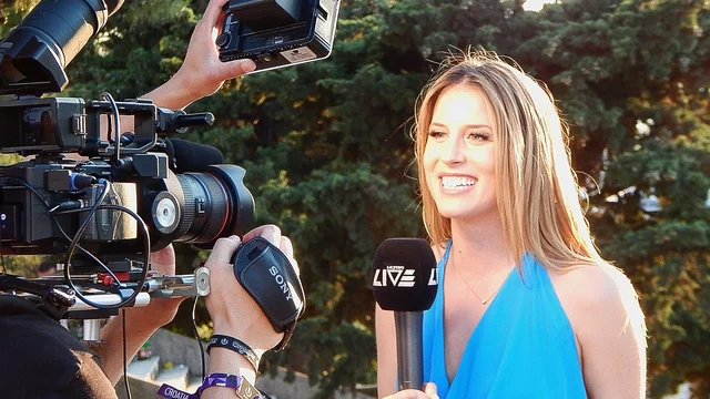Eine lächelnde Frau in einem blauen Kleid steht vor einer Kamera und spricht in ein Mikrofon. Im Hintergrund sind Bäume zu sehen. Die Szene vermittelt eine Atmosphäre von einem Live-Interview oder einer Berichterstattung.