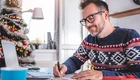 Ein Mann mit Brille sitzt an einem Schreibtisch und schreibt auf einen Kalender. Er trägt einen gestrickten Pullover mit Weihnachtsmuster. Im Hintergrund ist ein geschmückter Weihnachtsbaum zu sehen. Der Raum hat große Fenster und wirkt hell und einladend.