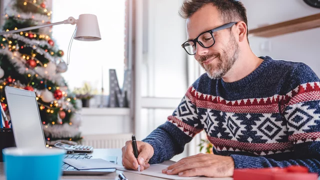 Ein Mann mit Brille sitzt an einem Schreibtisch und schreibt auf einen Kalender. Er trägt einen gestrickten Pullover mit Weihnachtsmuster. Im Hintergrund ist ein geschmückter Weihnachtsbaum zu sehen. Der Raum hat große Fenster und wirkt hell und einladend.