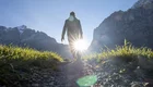 Eine Person geht auf einem Weg durch eine grüne Wiese, mit Bergen im Hintergrund. Die Sonne scheint hell hinter der Person, wodurch sie als Silhouette hervorsticht. Die Szenerie vermittelt ein Gefühl von Freiheit und Naturverbundenheit.