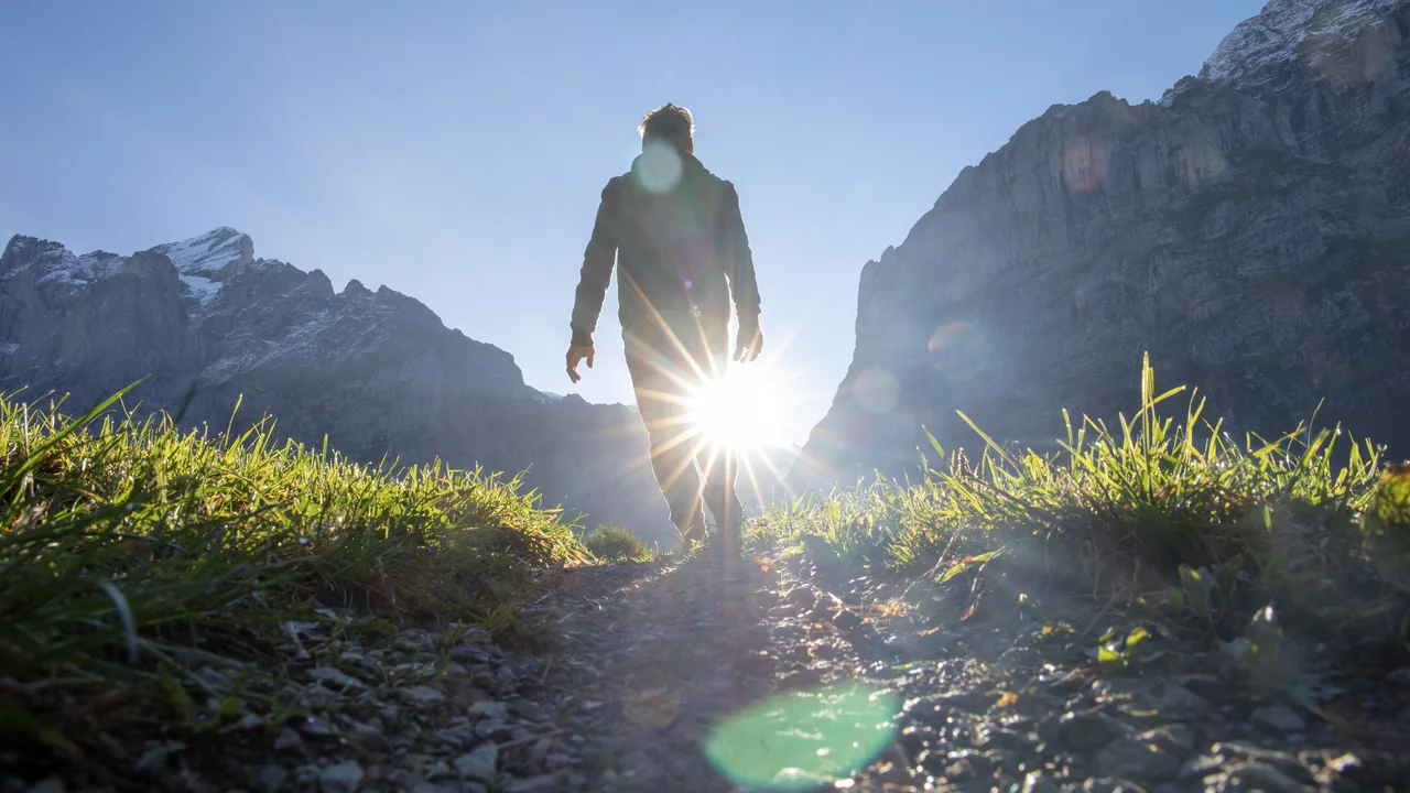 Eine Person geht auf einem Weg durch eine grüne Wiese, mit Bergen im Hintergrund. Die Sonne scheint hell hinter der Person, wodurch sie als Silhouette hervorsticht. Die Szenerie vermittelt ein Gefühl von Freiheit und Naturverbundenheit.