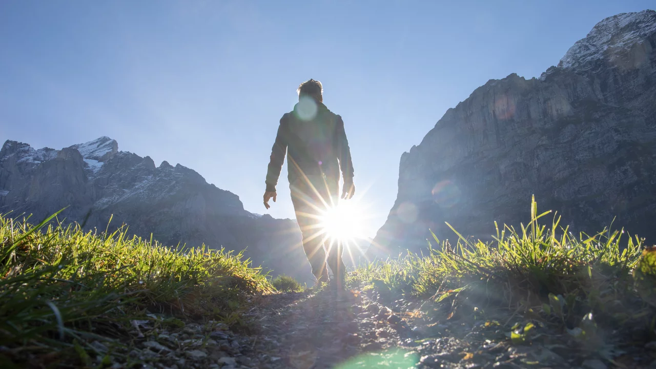 Eine Person geht auf einem Weg durch eine grüne Wiese, mit Bergen im Hintergrund. Die Sonne scheint hell hinter der Person, wodurch sie als Silhouette hervorsticht. Die Szenerie vermittelt ein Gefühl von Freiheit und Naturverbundenheit.
