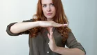 Eine Frau mit roten Locken steht vor einer neutralen Hintergrundfarbe und formt mit ihren Händen das Zeichen für Halt. Ihr Gesichtsausdruck ist ernst, und sie blickt direkt in die Kamera. Sie trägt ein graues Hemd und hat ihre Arme etwa auf Brusthöhe erhoben.