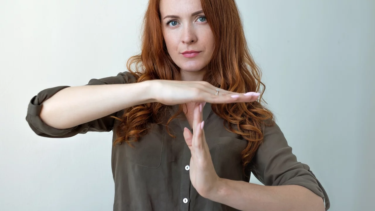 Eine Frau mit roten Locken steht vor einer neutralen Hintergrundfarbe und formt mit ihren Händen das Zeichen für Halt. Ihr Gesichtsausdruck ist ernst, und sie blickt direkt in die Kamera. Sie trägt ein graues Hemd und hat ihre Arme etwa auf Brusthöhe erhoben.