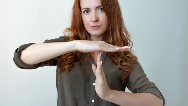 Eine Frau mit roten Locken steht vor einer neutralen Hintergrundfarbe und formt mit ihren Händen das Zeichen für Halt. Ihr Gesichtsausdruck ist ernst, und sie blickt direkt in die Kamera. Sie trägt ein graues Hemd und hat ihre Arme etwa auf Brusthöhe erhoben.
