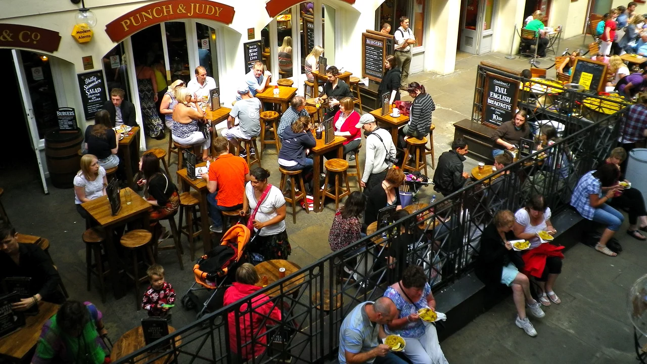 Eine belebte Außenfläche mit Menschen, die an hohen Tischen sitzen und essen oder trinken. Einige stehen, während andere an Tischen plaudern. Im Hintergrund sind ein paar Gastronomiebetriebe mit Schildern sichtbar. Die Atmosphäre wirkt gesellig und einladend.