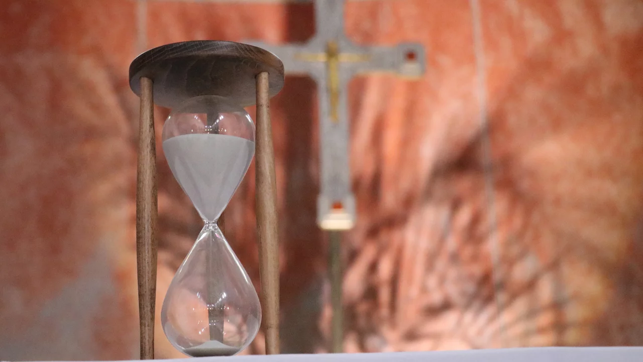 Eine Sanduhr steht auf einem Tisch, mit einer Holzstruktur oben und zwei gläsernen Kammern unten. Im Hintergrund ist ein Kreuz sichtbar. Die Sanduhr symbolisiert den Fluss der Zeit.