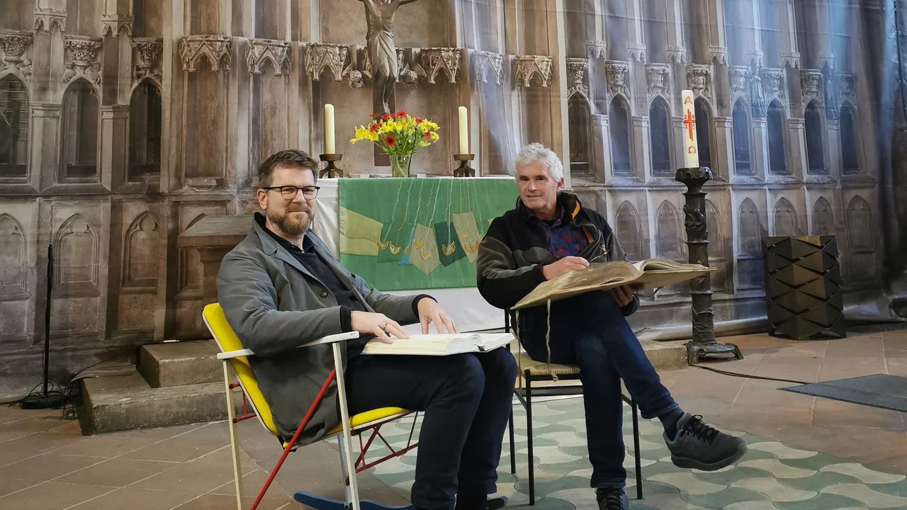 Zwei Männer sitzen in einer Kirche, umgeben von sakralen Elementen. Der erste Mann trägt eine Brille und hält ein Buch, während der zweite Mann in einem schwarzen Mantel ebenfalls ein Buch auf dem Schoß hat. Im Hintergrund ist eine Altarraum-Darstellung mit Blumen und einer Statue zu sehen.