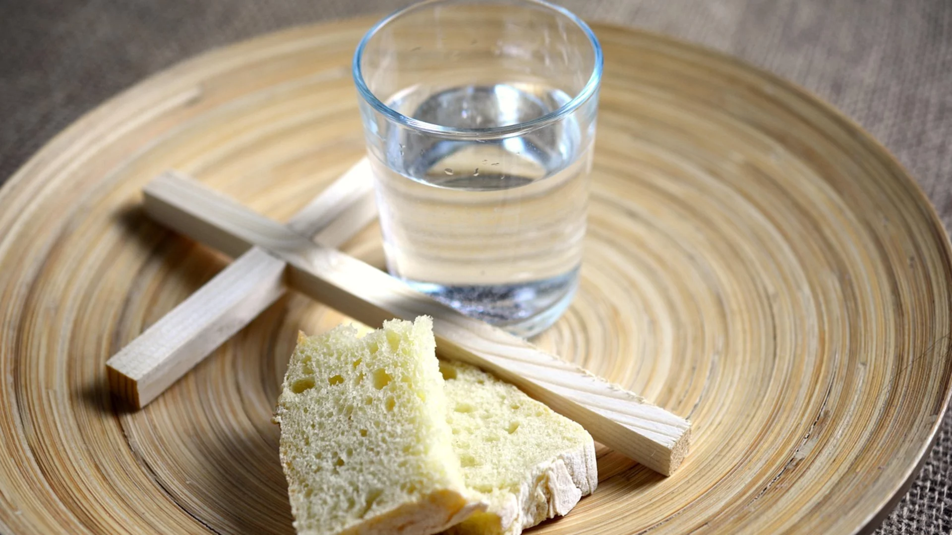 Auf einem runden Holztablett liegt ein Stück Brot und ein Glas Wasser. Zwei Holzstäbe sind über das Brot gelegt. Die Anordnung symbolisiert möglicherweise eine religiöse oder rituelle Bedeutung.