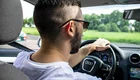 Ein junger Mann mit Sonnenbrille sitzt am Steuer eines Autos. Er trägt ein einfaches T-Shirt und hat eine modische Frisur. Im Hintergrund sind grüne Wiesen und ein leicht bewölkter Himmel sichtbar. Das Interieur des Autos ist modern und gut ausgestattet.