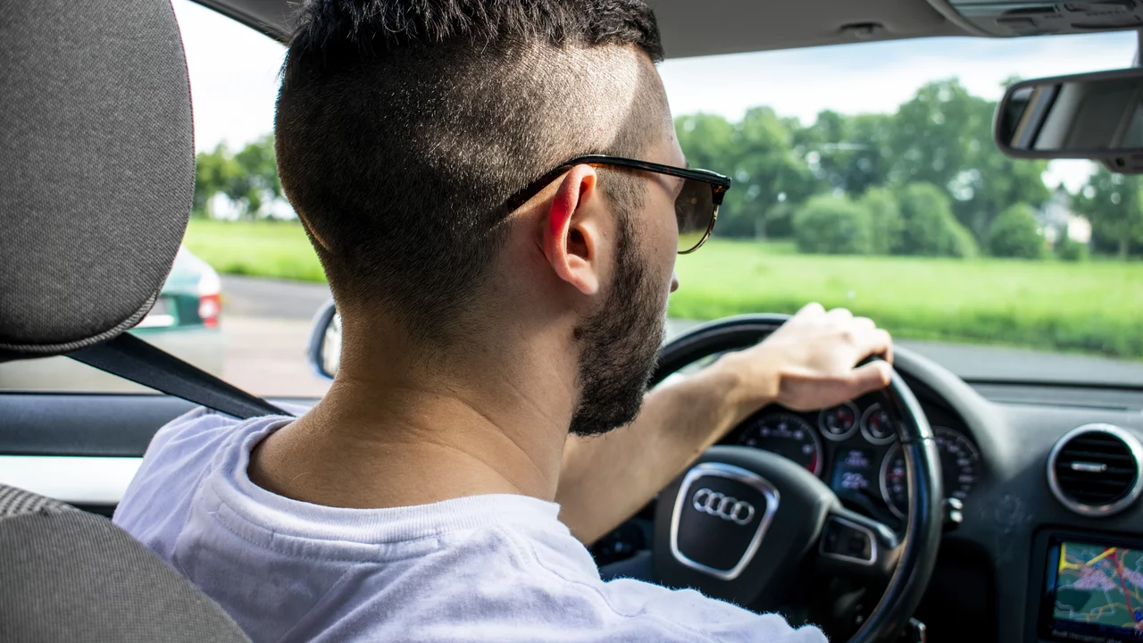 Ein junger Mann mit Sonnenbrille sitzt am Steuer eines Autos. Er trägt ein einfaches T-Shirt und hat eine modische Frisur. Im Hintergrund sind grüne Wiesen und ein leicht bewölkter Himmel sichtbar. Das Interieur des Autos ist modern und gut ausgestattet.
