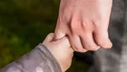 Eine erwachsene Hand hält die Hand eines Kindes. Die beiden Hände sind fest ineinander verschränkt, was eine Verbindung und Zuneigung zwischen den Personen symbolisiert. Der Hintergrund ist unscharf und zeigt eine grüne Wiese.