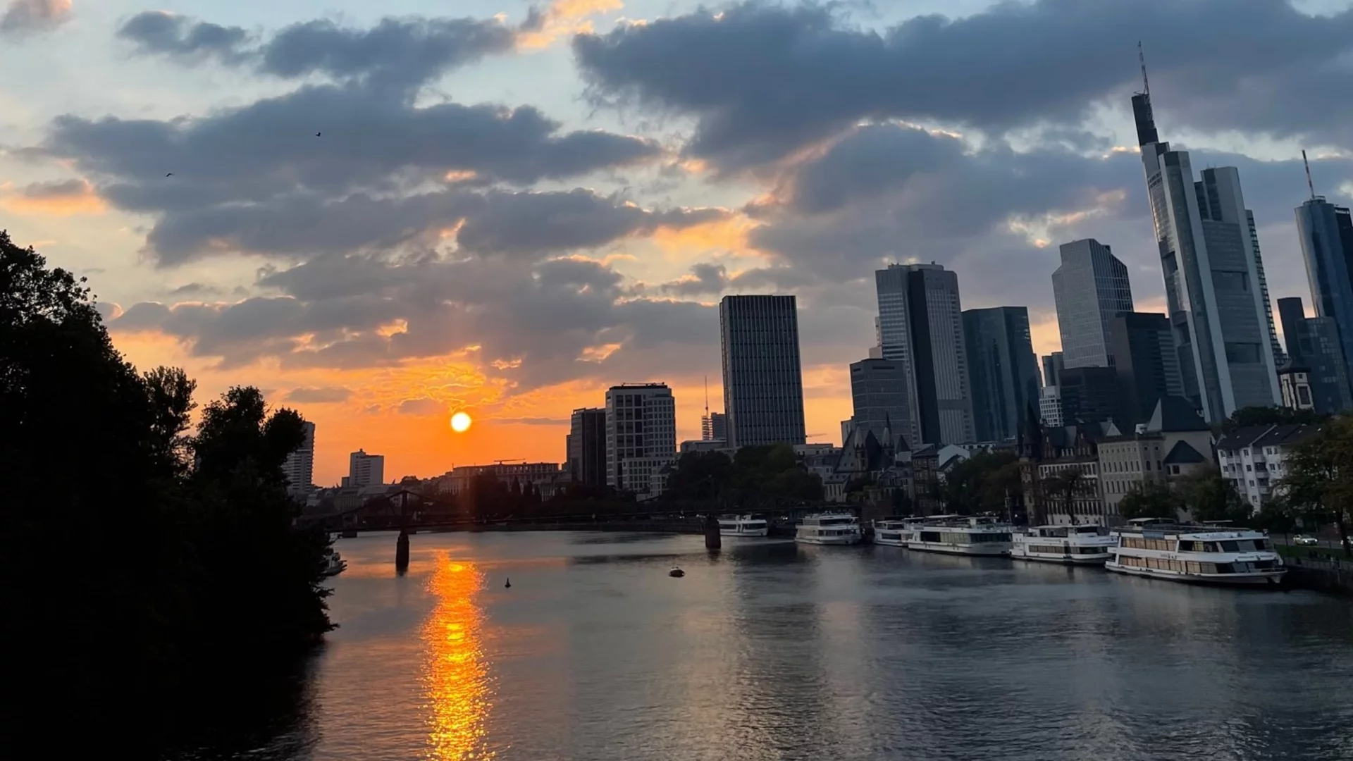 Sonnenuntergang über der Skyline von Frankfurt am Main. Die orangefarbenen und blauen Töne des Himmels spiegeln sich im Wasser des Flusses, während die Silhouetten der modernen Gebäude im Hintergrund erkennbar sind. Ein ruhiger, malerischer Moment in der Stadt.