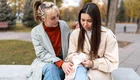 Zwei junge Frauen sitzen auf einer Bank in einem Park. Die erste Frau, mit einem hellen Mantel, zeigt Mitgefühl und legt ihre Hand auf den Arm der zweiten Frau, die traurig aussieht und in ihren Händen ein Taschentuch hält. Herbstliche Bäume sind im Hintergrund zu sehen.
