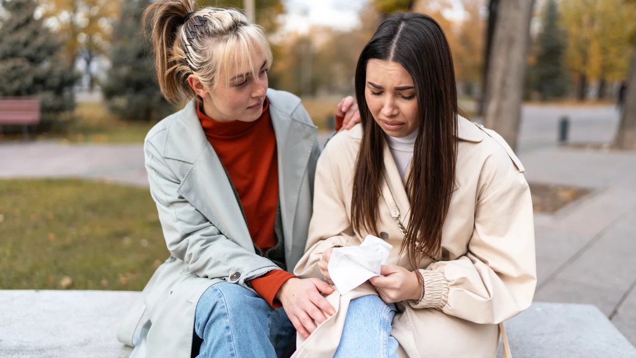Zwei junge Frauen sitzen auf einer Bank in einem Park. Die erste Frau, mit einem hellen Mantel, zeigt Mitgefühl und legt ihre Hand auf den Arm der zweiten Frau, die traurig aussieht und in ihren Händen ein Taschentuch hält. Herbstliche Bäume sind im Hintergrund zu sehen.