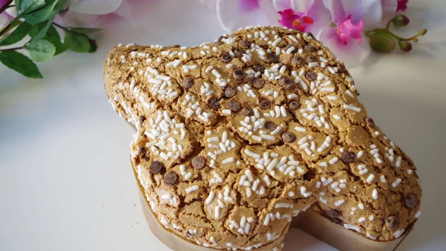 Ein großes, festliches Gebäck in Form eines Herzens, verziert mit Zuckerguss und Schokoladenstückchen. Das Gebäck hat eine rustikale, braune Oberfläche und ist auf einem einfachen Hintergrund platziert, umgeben von Blumen.