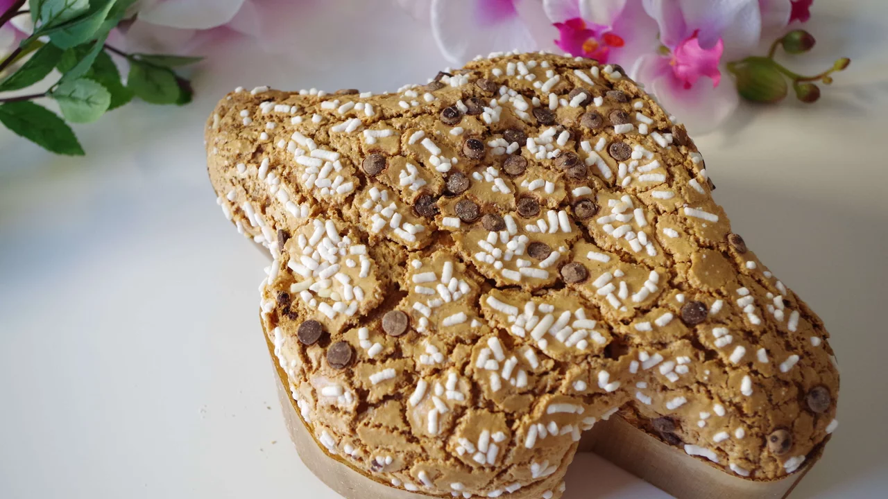 Ein großes, festliches Gebäck in Form eines Herzens, verziert mit Zuckerguss und Schokoladenstückchen. Das Gebäck hat eine rustikale, braune Oberfläche und ist auf einem einfachen Hintergrund platziert, umgeben von Blumen.