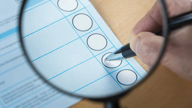 Eine Hand hält einen Stift und setzt ein Kreuz auf einen Wahlschein, der durch eine Lupe vergrößert wird. Der Fokus liegt auf dem markierten Kreis in der Wahl- oder Auswahlübersicht.